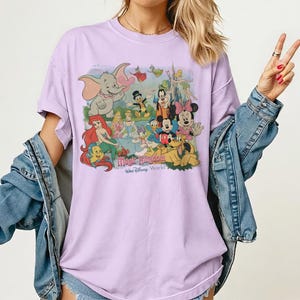 May include: A light purple t-shirt featuring a colorful graphic of Disney characters, including Dumbo, Mickey Mouse, and Ariel. The text "Magic Kingdom Walt Disney World" is printed below the characters. The shirt has short sleeves and a relaxed fit.