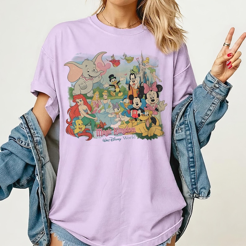 May include: A light purple t-shirt featuring a colorful graphic of Disney characters, including Dumbo, Mickey Mouse, and Ariel. The text "Magic Kingdom Walt Disney World" is printed below the characters. The shirt has short sleeves and a relaxed fit.