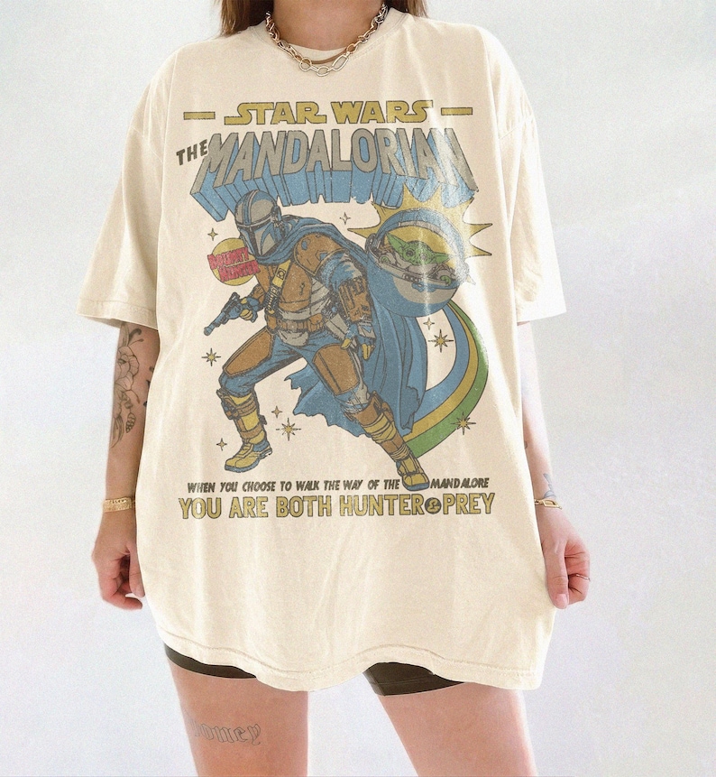 May include: Cream-colored t-shirt featuring a graphic of The Mandalorian from Star Wars. The design includes the Mandalorian, Grogu, and text that reads "STAR WARS THE MANDALORIAN" and "YOU ARE BOTH HUNTER PREY".