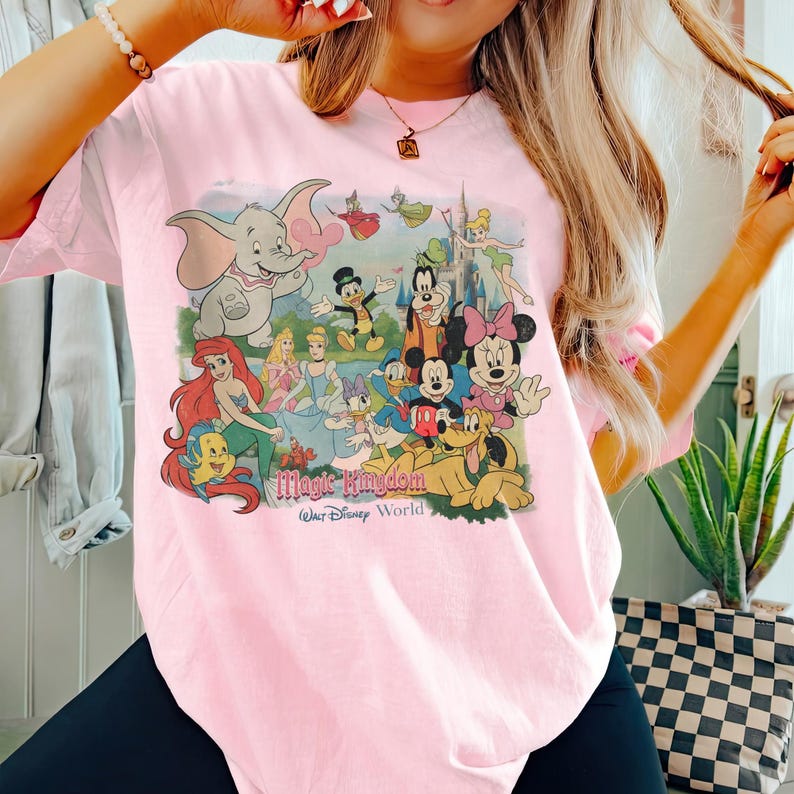 May include: A light pink t-shirt featuring a colorful graphic of Disney characters, including Dumbo, Ariel, Mickey and Minnie Mouse, and others. The words "Magic Kingdom Walt Disney World" are printed below the characters.
