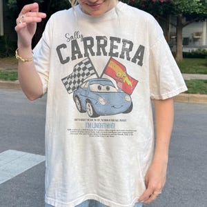 May include: Cream-colored t-shirt featuring a graphic of a blue car with the text "Sally Carrera" and "I'M LINGHTNING!". The design includes checkered and lightning bolt flags, evoking a racing theme.