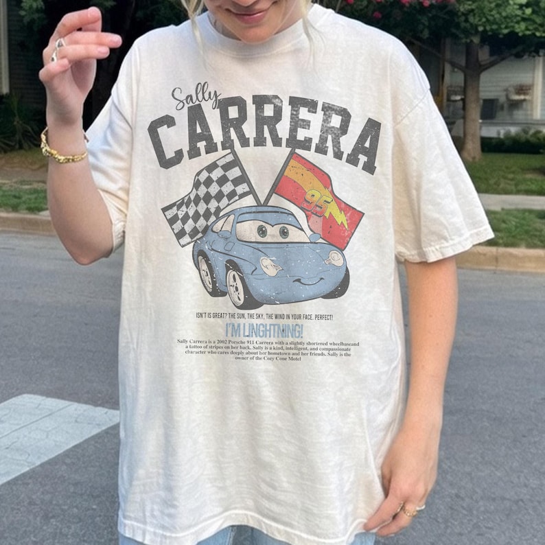 May include: Cream-colored t-shirt featuring a graphic of a blue car with the text "Sally Carrera" and "I'M LINGHTNING!". The design includes checkered and lightning bolt flags, evoking a racing theme.