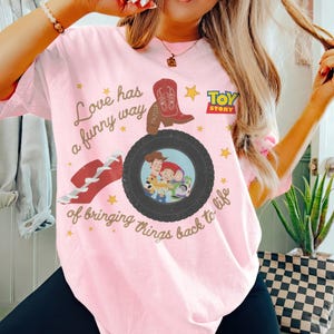 May include: A light pink t-shirt featuring a graphic print with the text "Love has a funny way of bringing things back to life," a red cowboy boot, a black tire, and the Toy Story logo. The tire contains an illustration of Woody, Jessie, and Buzz Lightyear.