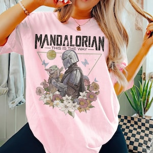 May include: Pink t-shirt featuring a graphic of The Mandalorian and Grogu surrounded by flowers and butterflies. The text "MANDALORIAN - THIS IS THE WAY" is printed across the top in black.