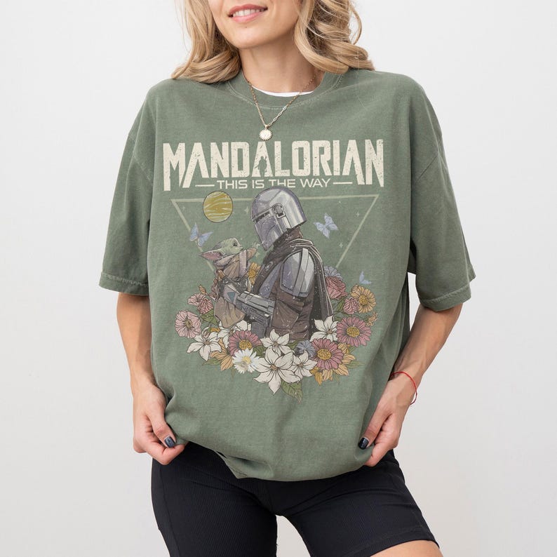 May include: Olive green t-shirt featuring a graphic of The Mandalorian and Grogu surrounded by flowers and butterflies. The text "MANDALORIAN - THIS IS THE WAY" is printed across the top. The shirt has short sleeves and a relaxed fit.