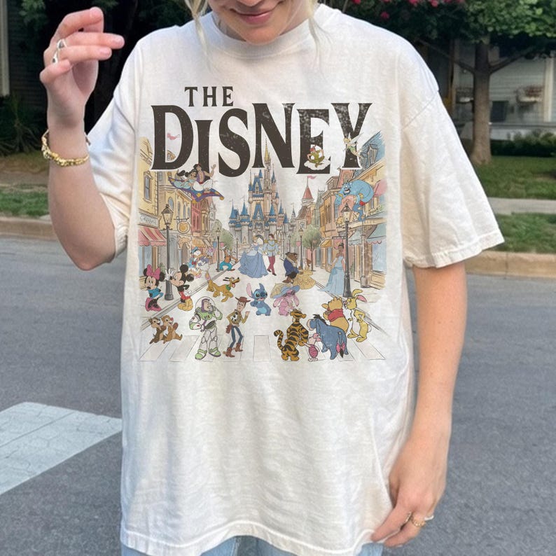 May include: Cream-coloured t-shirt with the text "THE DISNEY" above a colourful illustration of Disney characters and a street scene. The graphic includes Mickey Mouse, Winnie the Pooh, and other beloved characters.