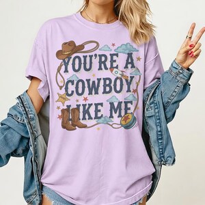 Puede incluir: Una camiseta de color morado claro con un estampado gr&aacute;fico que incluye el texto "YOU'RE A COWBOY LIKE ME" en letras azul oscuro. El dise&ntilde;o muestra un sombrero de vaquero marr&oacute;n, un lazo, botas vaqueras marrones, un cohete, una estrella y un yo-yo azul.