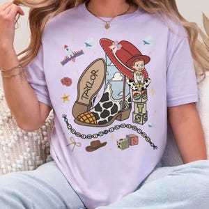 May include: A light purple t-shirt featuring a graphic print of a cow-print cowboy boot with the name TAYLOR, a red cowboy hat, a cartoon character, alphabet blocks, a rocket ship, and a beaded string with the text I KNEW IT WHEN YOU.