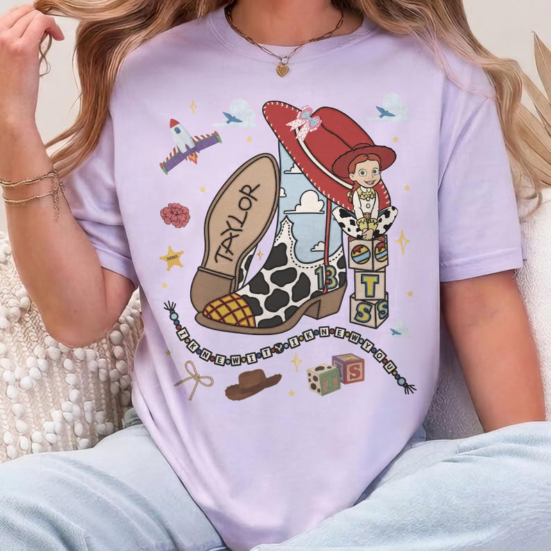 May include: A light purple t-shirt featuring a graphic print of a cow-print cowboy boot with the name TAYLOR, a red cowboy hat, a cartoon character, alphabet blocks, a rocket ship, and a beaded string with the text I KNEW IT WHEN YOU.