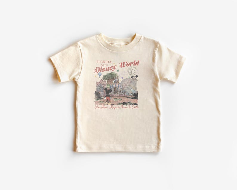 Puede incluir: Camiseta color crema con un gr&aacute;fico de Disney World de estilo vintage. El dise&ntilde;o incluye el texto "Florida Est. 1971 Disney World" y una ilustraci&oacute;n de Mickey Mouse y otros elementos del parque. La camiseta tiene mangas cortas y cuello redondo.