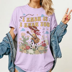 May include: A light purple t-shirt featuring a graphic of a cowgirl playing an acoustic guitar in a desert landscape with cacti and flowers. The text "I KNEW IT I KNEW YOU" is printed in a retro yellow font above the character, surrounded by small stars, musical notes, and clouds.