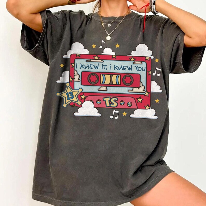 May include: A charcoal gray t-shirt featuring a graphic of a red cassette tape with the text "I KNEW IT, I KNEW YOU" and "TS". The design includes white clouds, yellow stars, and musical notes, with a yellow star badge labeled with the number 13.