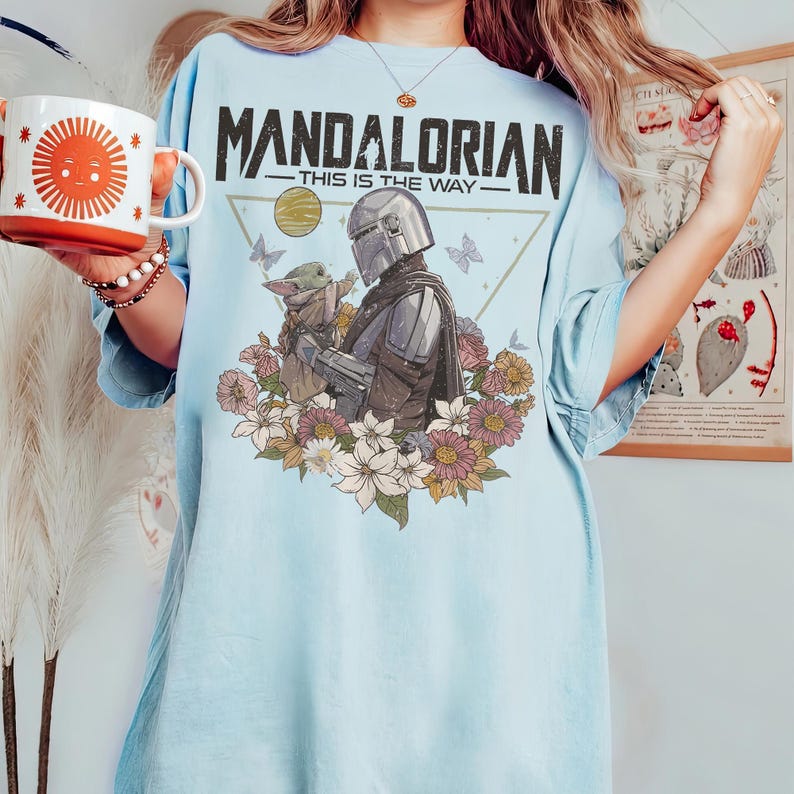 May include: Light blue t-shirt featuring a graphic of The Mandalorian and Grogu surrounded by flowers. The text "MANDALORIAN - THIS IS THE WAY" is printed across the top. A white mug with an orange sun design is held in the left hand.