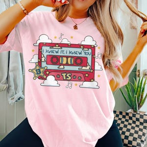 May include: A light pink t-shirt featuring a graphic of a red cassette tape with the text I KNEW IT, I KNEW YOU. The design includes a yellow star with the number 13, the letters TS, white clouds, musical notes, and small yellow stars.