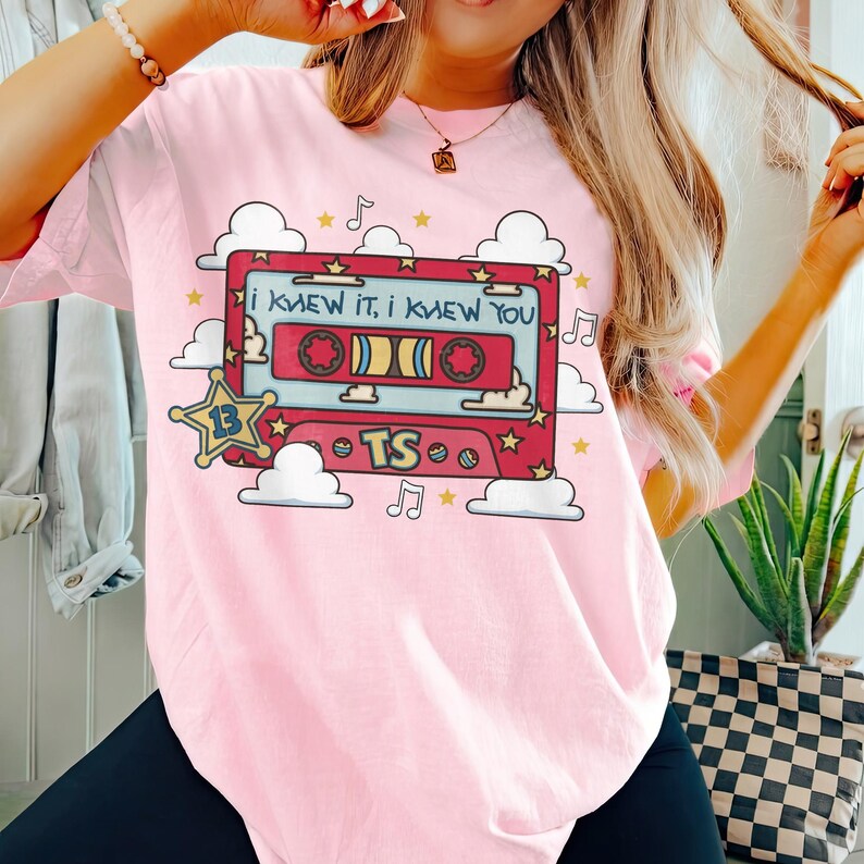 May include: A light pink t-shirt featuring a graphic of a red cassette tape with the text I KNEW IT, I KNEW YOU. The design includes a yellow star with the number 13, the letters TS, white clouds, musical notes, and small yellow stars.