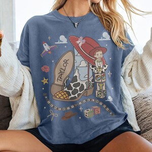 May include: A blue t-shirt featuring a graphic print of a cow-print cowboy boot with the name TAYLOR on the sole, a red cowboy hat, a cartoon character, building blocks with the letters T S, a rocket, and the text I KNEW IT I KNEW YOU.