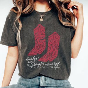 K&ouml;nnte beinhalten: Ein anthrazitfarbenes Kurzarm-T-Shirt mit einer Grafik aus zwei roten Cowboystiefeln, die mit Herzen, Sternen und der Zahl 13 verziert sind. Unter den Stiefeln steht in wei&szlig;er Schreibschrift der Text: Love has ways of bringing things back to life.