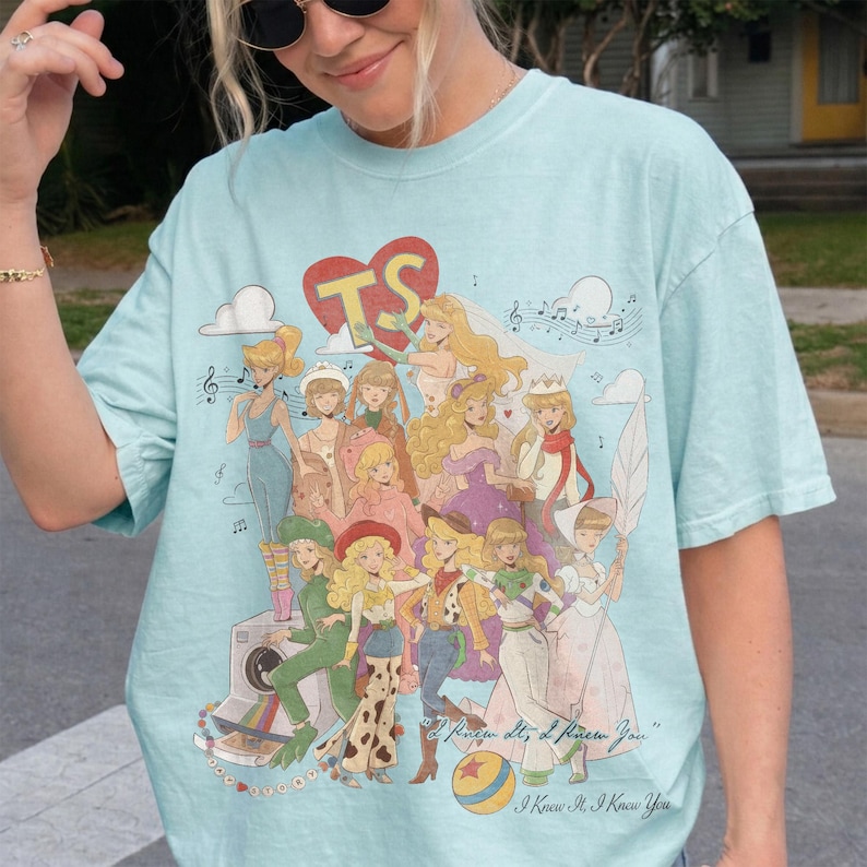 May include: A light blue t-shirt featuring a large graphic print of multiple blonde characters in various outfits, a red heart with the letters TS, musical notes, clouds, a camera, and a ball. The text reads "I Knew It, I Knew You".