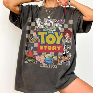 May include: Dark gray t-shirt featuring a colorful graphic of Toy Story characters, including Buzz Lightyear, Woody, and Jessie. The design includes the words "TOY STORY" in yellow and red, with "EST.1995" below. The shirt has a vintage, worn look.