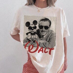 May include: Cream-colored t-shirt featuring a vintage-style graphic of Mickey Mouse wearing sunglasses alongside Walt Disney. The word "Walt" is printed in red script across the bottom of the graphic. The shirt has a classic crew neck.