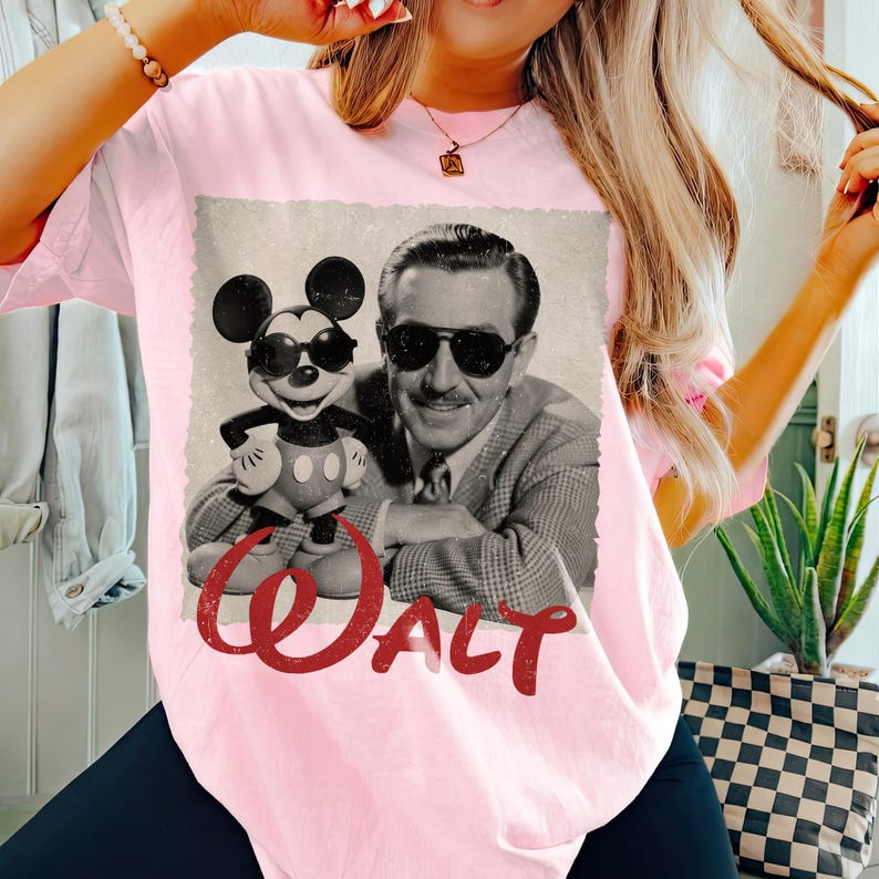 May include: Pink t-shirt featuring a vintage-style graphic of Mickey Mouse wearing sunglasses alongside Walt Disney, with the word "Walt" in red script. The image has a distressed, aged look, evoking nostalgia.