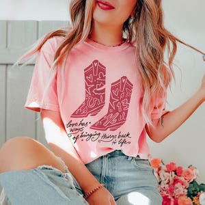 K&ouml;nnte beinhalten: Ein hellrosa T-Shirt mit kurzen &Auml;rmeln und einem dunkelrosa Aufdruck von zwei Cowboystiefeln, die mit Herzen, Sternen und der Zahl 13 verziert sind. Unter den Stiefeln steht der Text Love has ways of bringing things back to life.