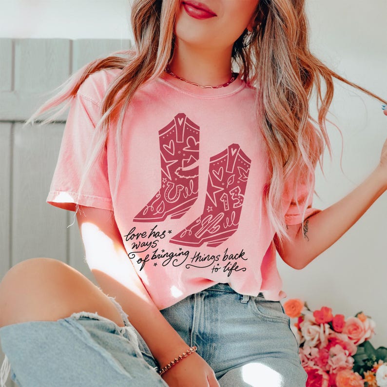 K&ouml;nnte beinhalten: Ein hellrosa T-Shirt mit kurzen &Auml;rmeln und einem dunkelrosa Aufdruck von zwei Cowboystiefeln, die mit Herzen, Sternen und der Zahl 13 verziert sind. Unter den Stiefeln steht der Text Love has ways of bringing things back to life.