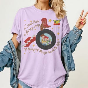 May include: A light purple t-shirt featuring a graphic print with the text "Love has a funny way of bringing things back to life," a red cowboy boot, a red hat, yellow stars, the Toy Story logo, and a tire containing Woody, Jessie, and Buzz Lightyear.
