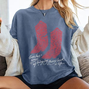 K&ouml;nnte beinhalten: Ein schieferblaues T-Shirt mit kurzen &Auml;rmeln und einer Grafik von zwei roten Cowboystiefeln, die mit Herzen und Sternen verziert sind. Unter den Stiefeln steht in wei&szlig;er Schreibschrift der Text: Love has ways of bringing things back to life.