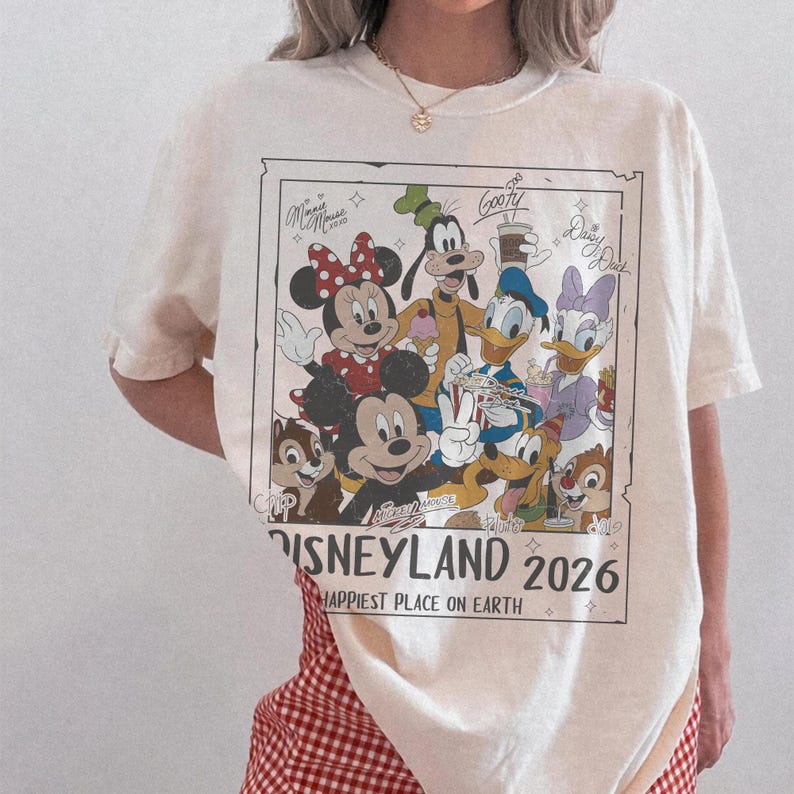 May include: Cream-colored t-shirt featuring a Disneyland 2026 graphic with Mickey Mouse, Minnie Mouse, Goofy, Donald Duck, Daisy Duck, Chip, and Dale. The text reads "Happiest Place on Earth."