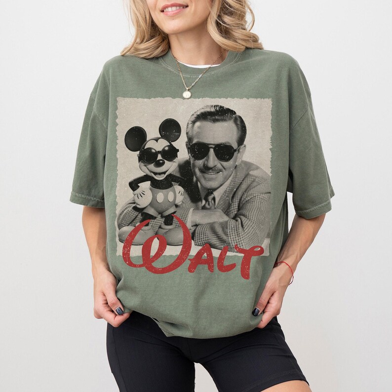 May include: Olive green t-shirt featuring a vintage graphic of Walt Disney with Mickey Mouse wearing sunglasses. The word "Walt" is printed in red script across the bottom of the graphic. The shirt is a relaxed fit.