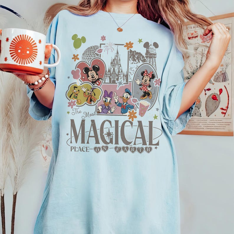 May include: Light blue t-shirt featuring a colorful graphic with Mickey and Minnie Mouse, Donald and Daisy Duck, and Goofy. The text reads "The Most Magical Place on Earth." A red and white mug with a sun design is held in the hand.