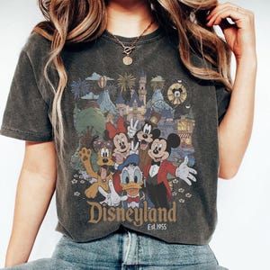 May include: Dark gray t-shirt featuring a colorful graphic of Disney characters, including Mickey Mouse, Minnie Mouse, and Donald Duck, in front of a castle and fireworks. The word "Disneyland" is printed below the graphic, with "Est.1955" underneath.