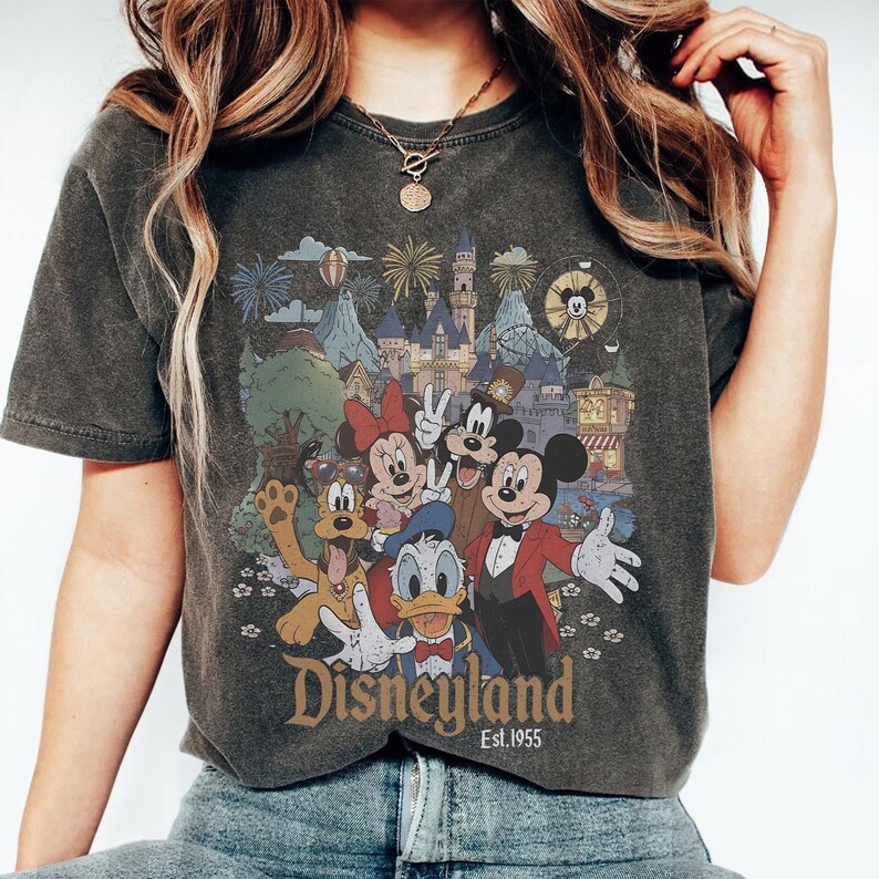 May include: Dark gray t-shirt featuring a colorful graphic of Disney characters, including Mickey Mouse, Minnie Mouse, and Donald Duck, in front of a castle and fireworks. The word "Disneyland" is printed below the graphic, with "Est.1955" underneath.