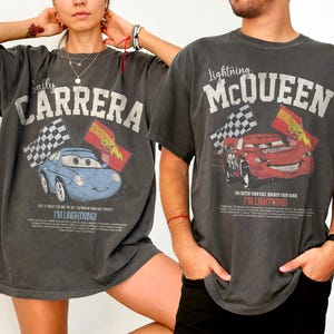 May include: Two charcoal gray t-shirts featuring graphics from the movie Cars. One shirt displays "Sally Carrera" with a blue car and checkered flags. The other shirt shows "Lightning McQueen" with a red car and checkered flags. Both shirts have text that reads "I'M LIGHTNING!"