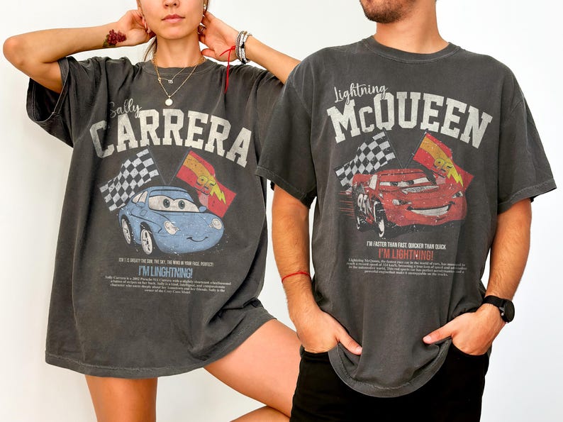 May include: Two charcoal gray t-shirts featuring graphics from the movie Cars. One shirt displays "Sally Carrera" with a blue car and checkered flags. The other shirt shows "Lightning McQueen" with a red car and checkered flags. Both shirts have text that reads "I'M LIGHTNING!"