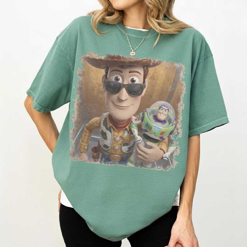 May include: A sage green short-sleeve t-shirt featuring a graphic print of Woody wearing sunglasses and holding Buzz Lightyear. The design is framed within a distressed, rectangular border, showcasing the characters from the Toy Story franchise.