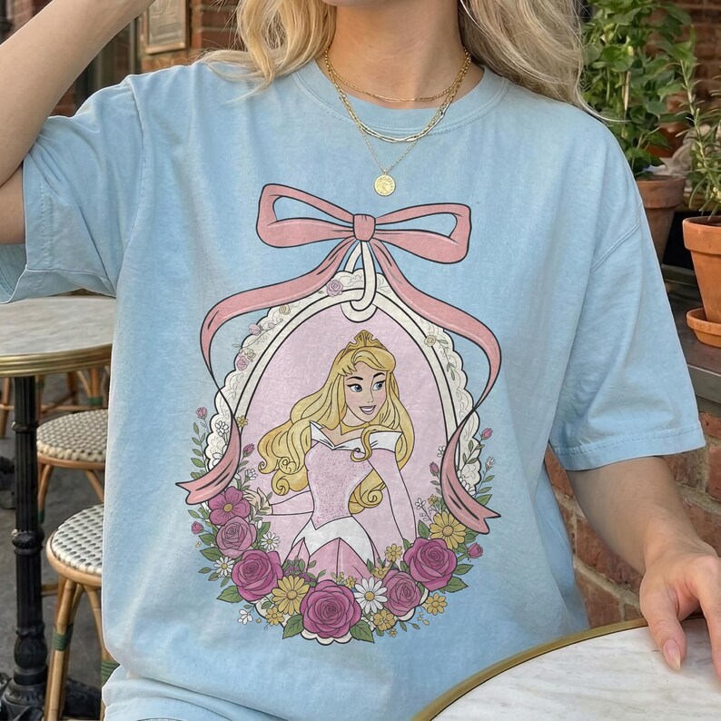 K&ouml;nnte beinhalten: Hellblaues T-Shirt mit einer gerahmten Illustration einer Prinzessin in einem rosa Kleid, umgeben von rosa Rosen und gelben Blumen. &Uuml;ber dem Rahmen befindet sich eine rosa Schleife. Freizeitkleidung.