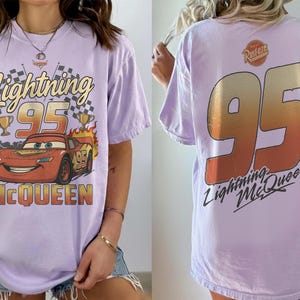 K&ouml;nnte beinhalten: Lavendelfarbenes T-Shirt mit Lightning McQueen-Design. Auf der Vorderseite steht "Lightning 95 McQueen" mit Rennwagen, Flaggen und Troph&auml;en. Auf der R&uuml;ckseite steht eine gro&szlig;e "95" mit "Lightning McQueen" darunter. Ein l&auml;ssiges Shirt.
