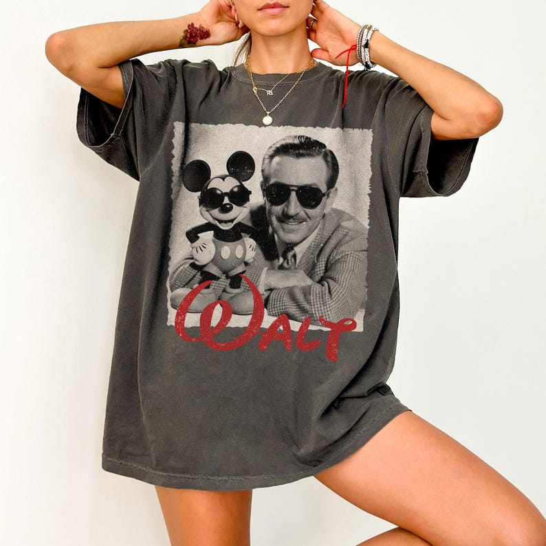 May include: Dark gray t-shirt featuring a black and white image of Mickey Mouse wearing sunglasses and Walt Disney. The word "Walt" is printed in red script below the image. The shirt has short sleeves and a relaxed fit.