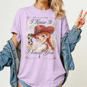 May include: A light purple t-shirt featuring a graphic of a cartoon cowgirl with red hair and a brown hat. The text reads EST 2026, I Knew It, and I Knew You. The design includes a rope border, stars, and horseshoes, with a blue denim jacket layered over.