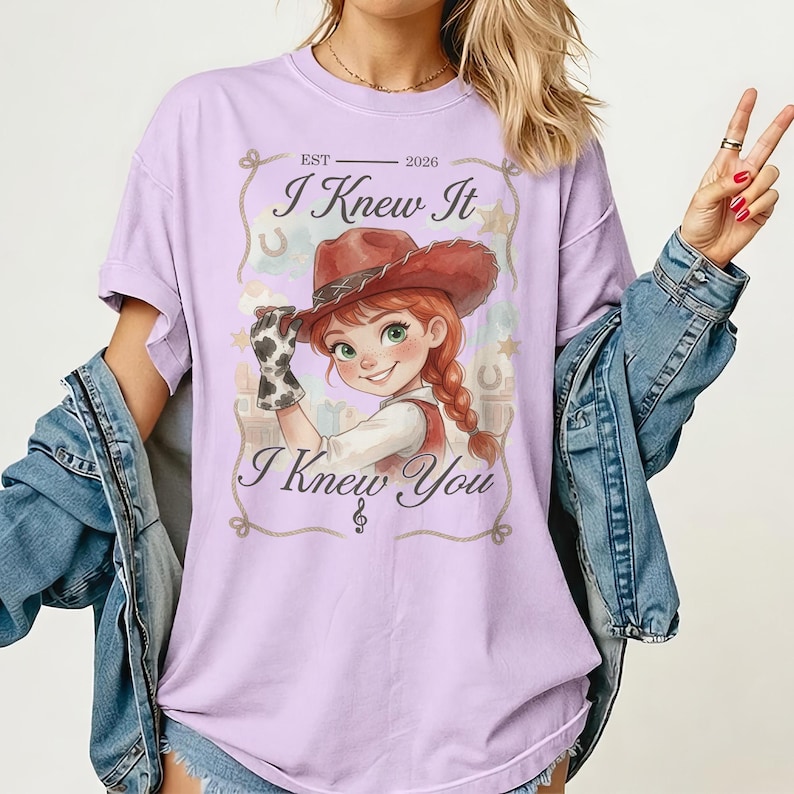 May include: A light purple t-shirt featuring a graphic of a cartoon cowgirl with red hair and a brown hat. The text reads EST 2026, I Knew It, and I Knew You. The design includes a rope border, stars, and horseshoes, with a blue denim jacket layered over.