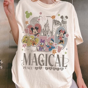May include: Cream-colored t-shirt featuring a colorful graphic design with Mickey and Minnie Mouse, Donald Duck, Daisy Duck, and Goofy. The text reads "The Most Magical Place on Earth" in a whimsical font. Floral and heart accents complete the design.