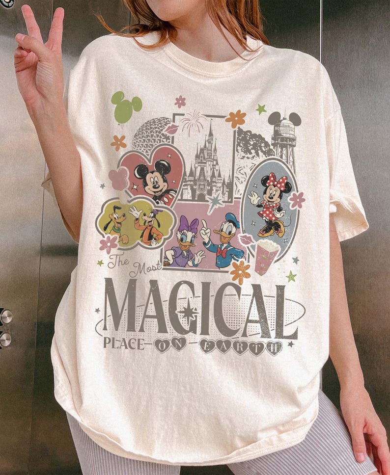 May include: Cream-colored t-shirt featuring a colorful graphic design with Mickey and Minnie Mouse, Donald Duck, Daisy Duck, and Goofy. The text reads "The Most Magical Place on Earth" in a whimsical font. Floral and heart accents complete the design.