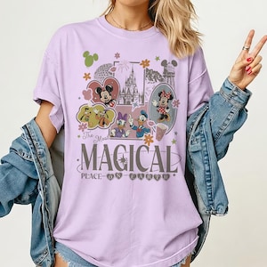 May include: A lilac-colored t-shirt featuring a graphic design with Disney characters, including Mickey and Minnie Mouse, Donald and Daisy Duck, and Goofy. The design includes the text "The Most Magical Place on Earth" with floral and star accents.