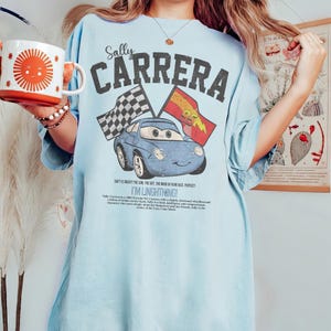 May include: Light blue t-shirt featuring a cartoon car with the text "Sally Carrera" and "I'M LINGHTNING!". The car is depicted with checkered and "95" flags. A mug with a sun design is held in the image.