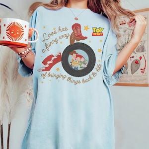 May include: A light blue t-shirt featuring a graphic print with the text "Love has a funny way of bringing things back to life," a red cowboy boot, a black tire containing an illustration of Woody, Jessie, and Buzz Lightyear, and a Toy Story logo.