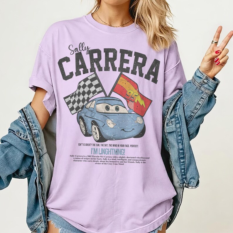 May include: A lilac-colored t-shirt featuring a graphic of Sally Carrera from the movie Cars. The design includes the text "Sally Carrera" above the car image, and "I'M LINGHTNING!" below. Two flags are also included in the design.