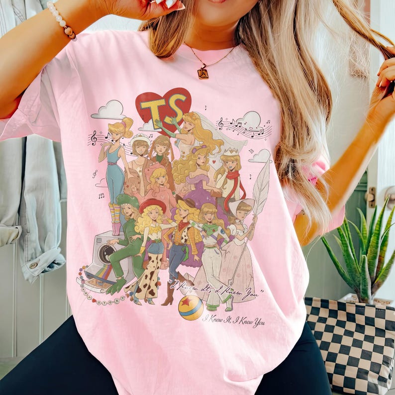 May include: A light pink t-shirt featuring a graphic print of various animated female characters in diverse outfits, a large red heart with the letters TS, musical notes, a camera, and a ball. The text reads, I Knew It, I Knew You.