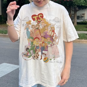 May include: An off-white short-sleeve t-shirt featuring a large graphic print of multiple illustrated female characters, a red heart with the letters TS, a camera, and a ball. The text I Knew It, I Knew You is printed in a cursive script at the bottom.